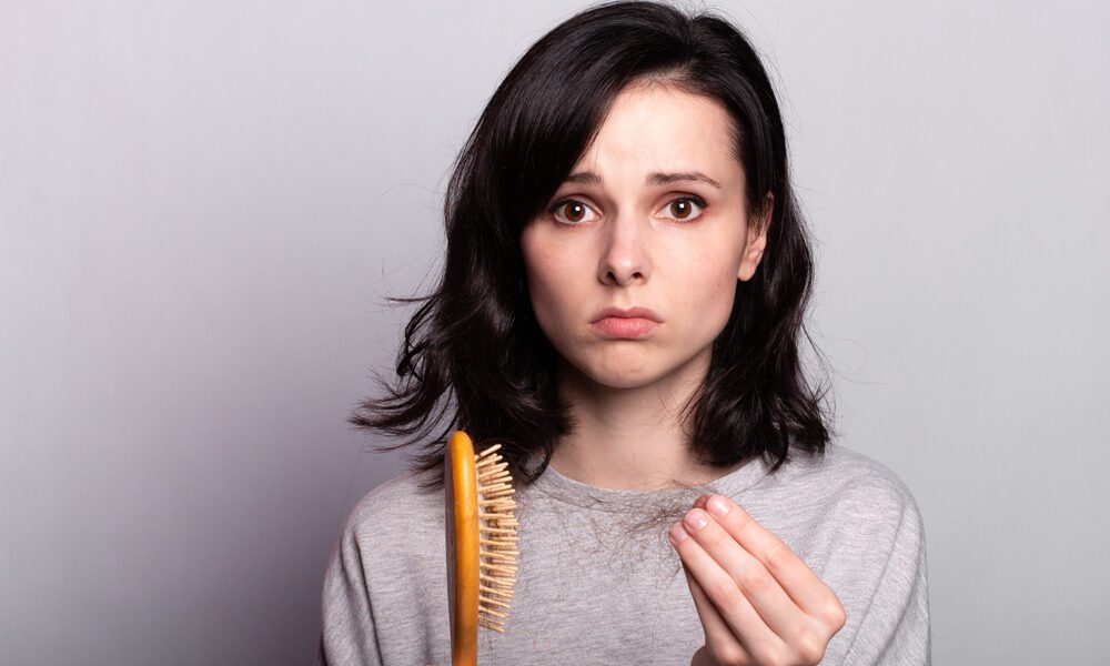 Em fundo cinza, mulher segura uma escova em uma mão e um tufo de cabelo na outra